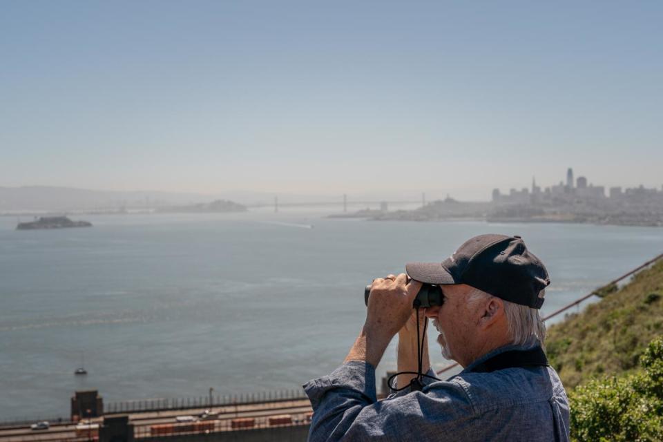 Bill Keener looks through binoculars at San Francisco Bay, with the city skyline in the background.