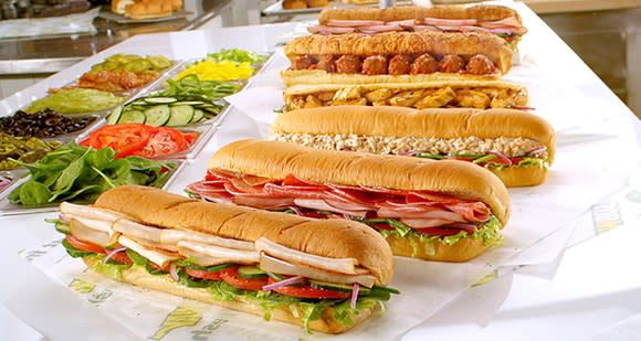 Six Subway sandwiches lined up on a countertop, with various vegetables displayed alongside.