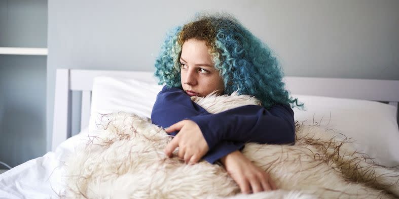 young woman with dyed blue hair looking sad whilst sat up in bed and gazing off into the distance