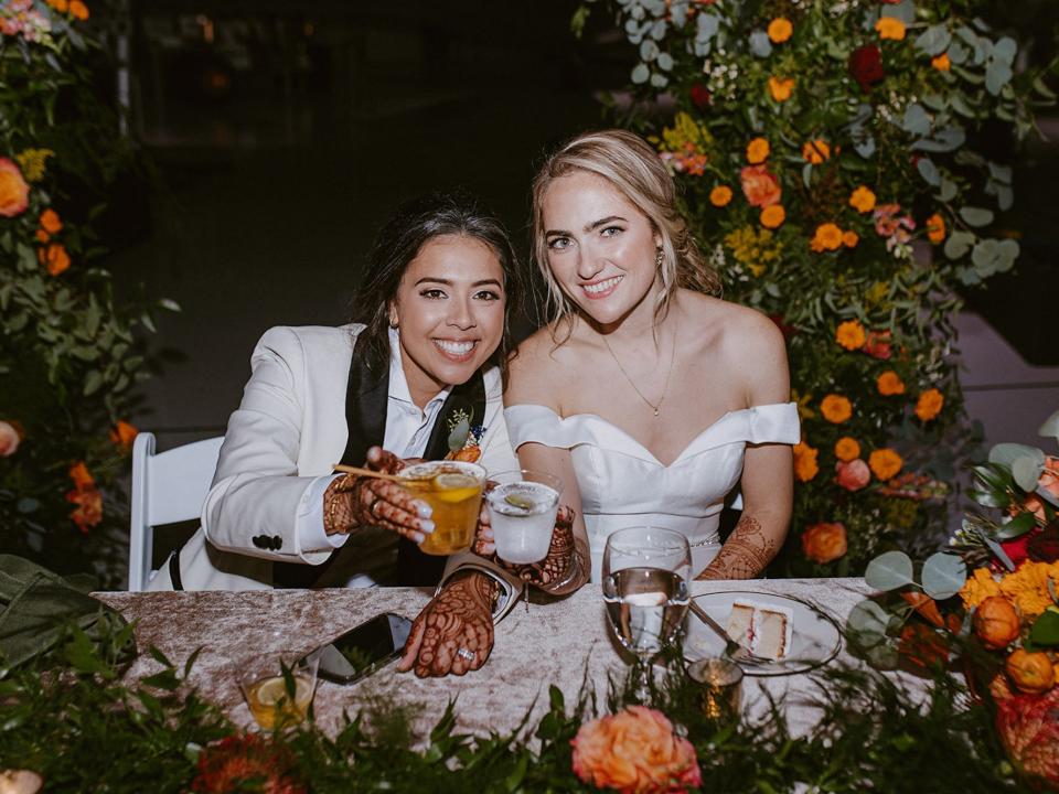 Two brides cheers their glasses at a table in front of flowers.