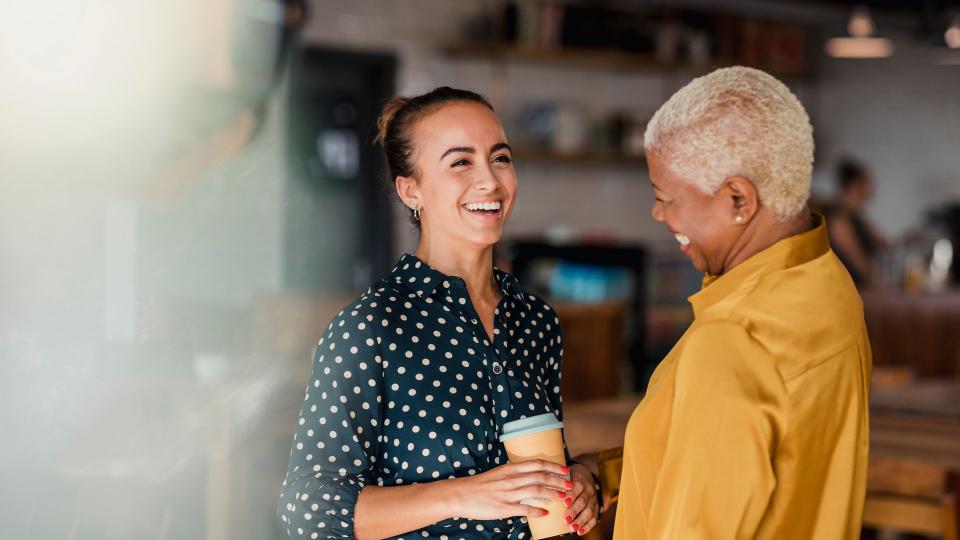 Two women colleagues laughing while standing in a cafe at their workplace. One of the women is holding a take out hot drink cup.