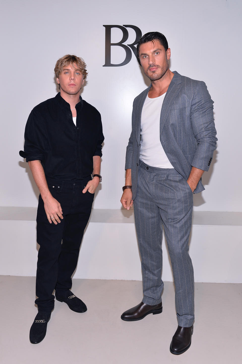 Two men posing at an event, one in a black outfit and the other in a pinstriped suit with a white shirt