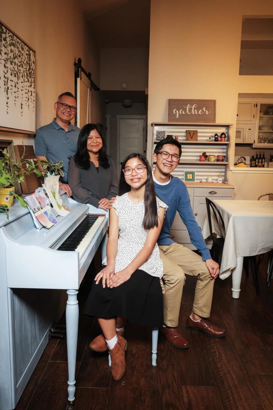 A girl sits on a piano bench with her brother. Her parents stand behind them and the white piano.