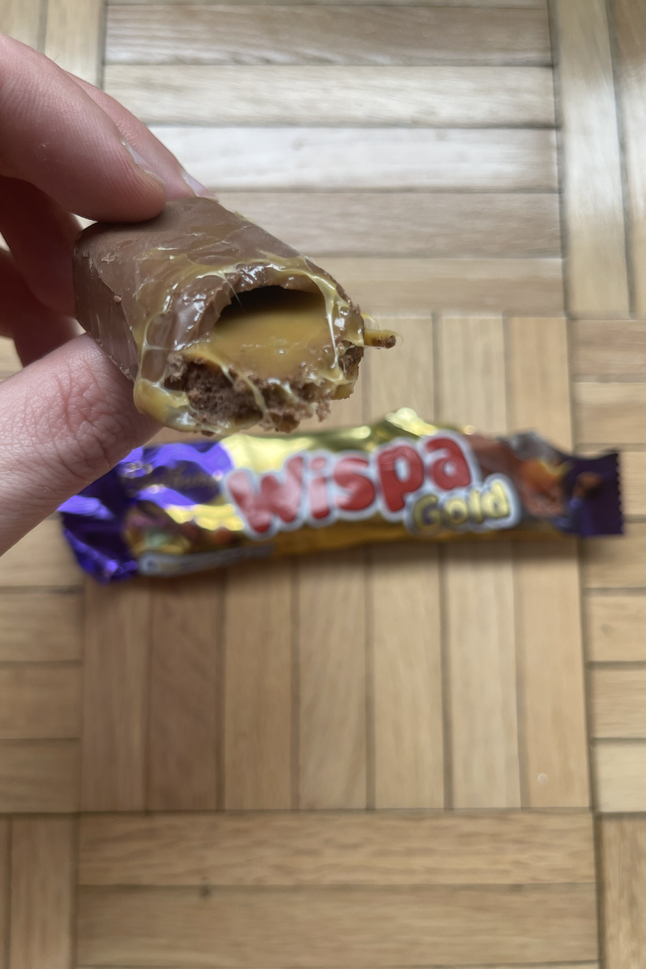 Hand holding a partially eaten Wispa Gold chocolate bar over a wooden floor with wrapper in the background