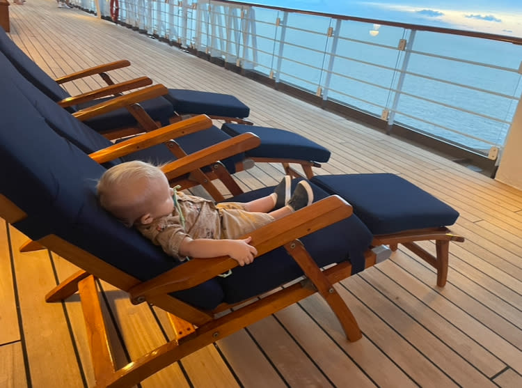 A child is lounging on a deck chair on a ship's deck, with a calm sea visible in the background