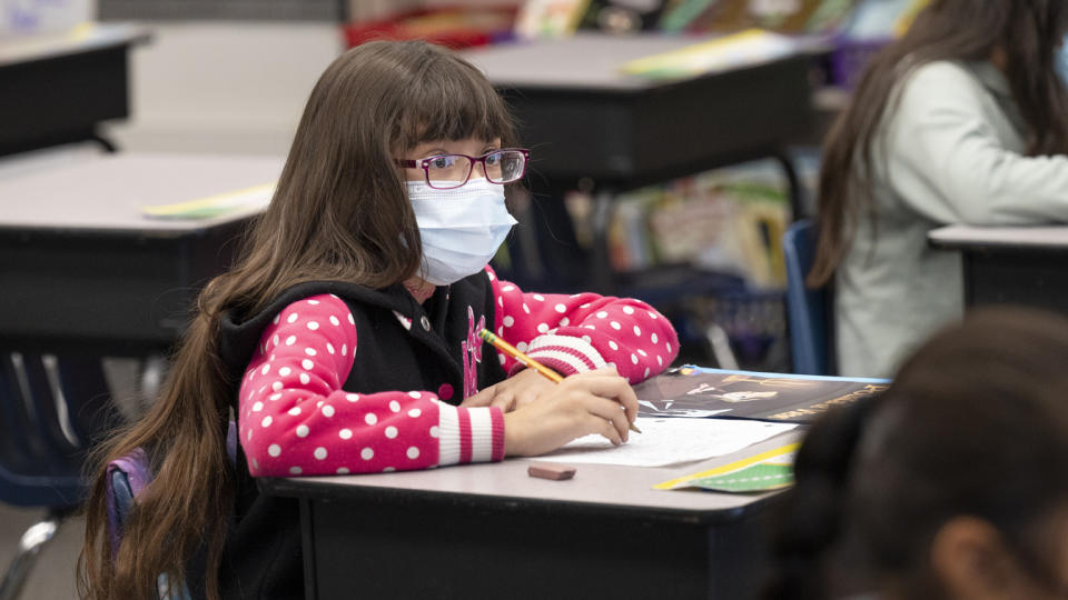 A student wearing a face mask holds a pencil while sitting at a school desk.
