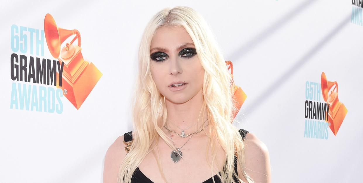 taylor momsen, a young woman stands looking at the camera, she has long blonde hair worn down in loose waves, heavy black eye makeup and wears a short black dress