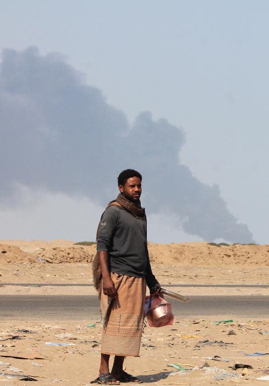 A Yemeni man carries kitchen utensils as he walks on a main road with smoke rising in the background from a strike allegedly targeting Huthi rebels on the outskirts in Yemen's second city of Aden on March 30, 2015