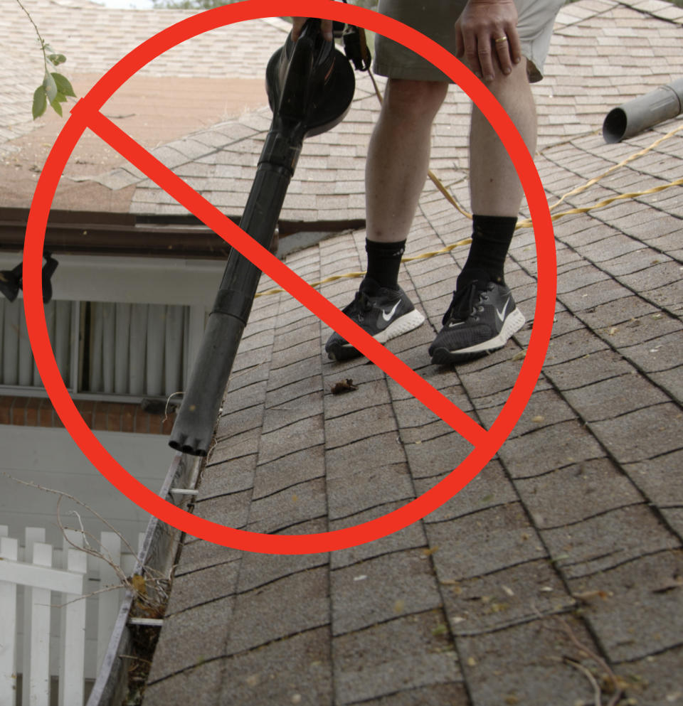 A person wearing shorts and sneakers is using a leaf blower to clear debris from a gutter on a shingled roof