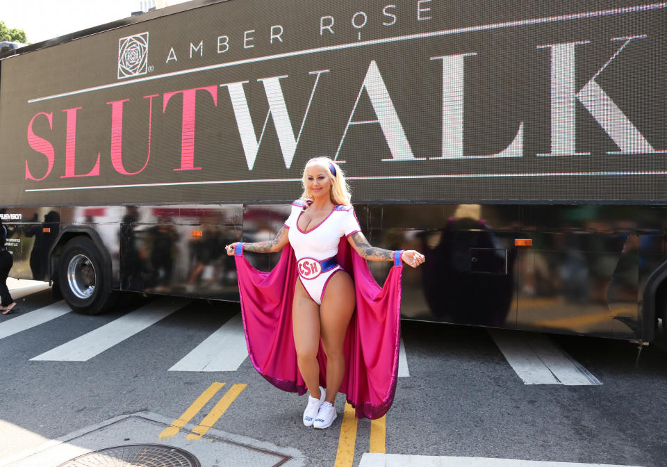 Amber Rose poses in front of a large "SlutWalk" banner, dressed in a superhero-style outfit with a cape bearing the initials "SR"