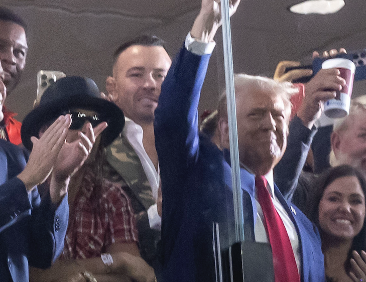 Donald Trump and Melania Trump smile and wave with a crowd of people, some taking photos, behind a glass barrier at an event