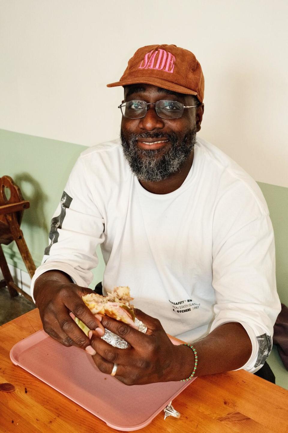 A vertical photo of Toks Shoyoye sitting at a booth Cafe Tropical, holding a sandwich on reopening day in Silver Lake