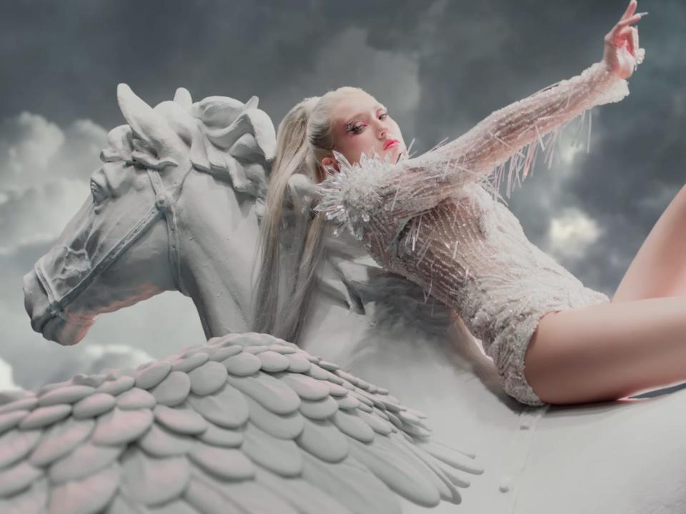 jeon somi in fast forward, wearing a white, sparkling unitard with spiky crystal shoulders, a long blonde ponytail, and laying across a statue of a pegasus