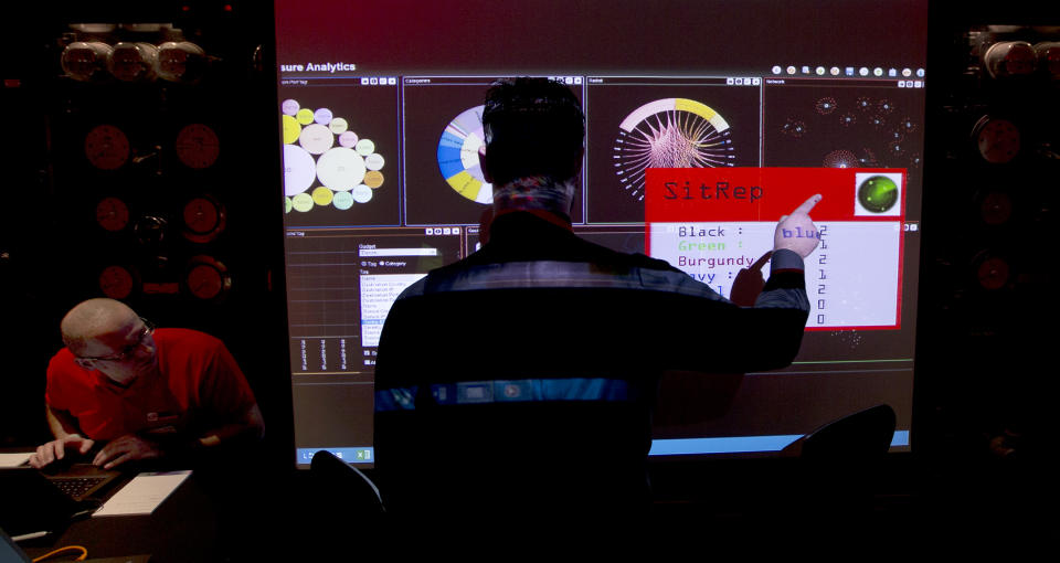 A representative of GCHQ points to a screen showing all the teams progress in completing the task during a mock cyberattack scenario with teams of amateur computer experts taking part and trying to fight this simulated attack in London, Friday, March, 14, 2014. GCHQ is the British Government's electronic intelligence service, they describe themselves as the technical partner to the intelligence and security services, MI6 and MI5. (AP Photo/Alastair Grant)