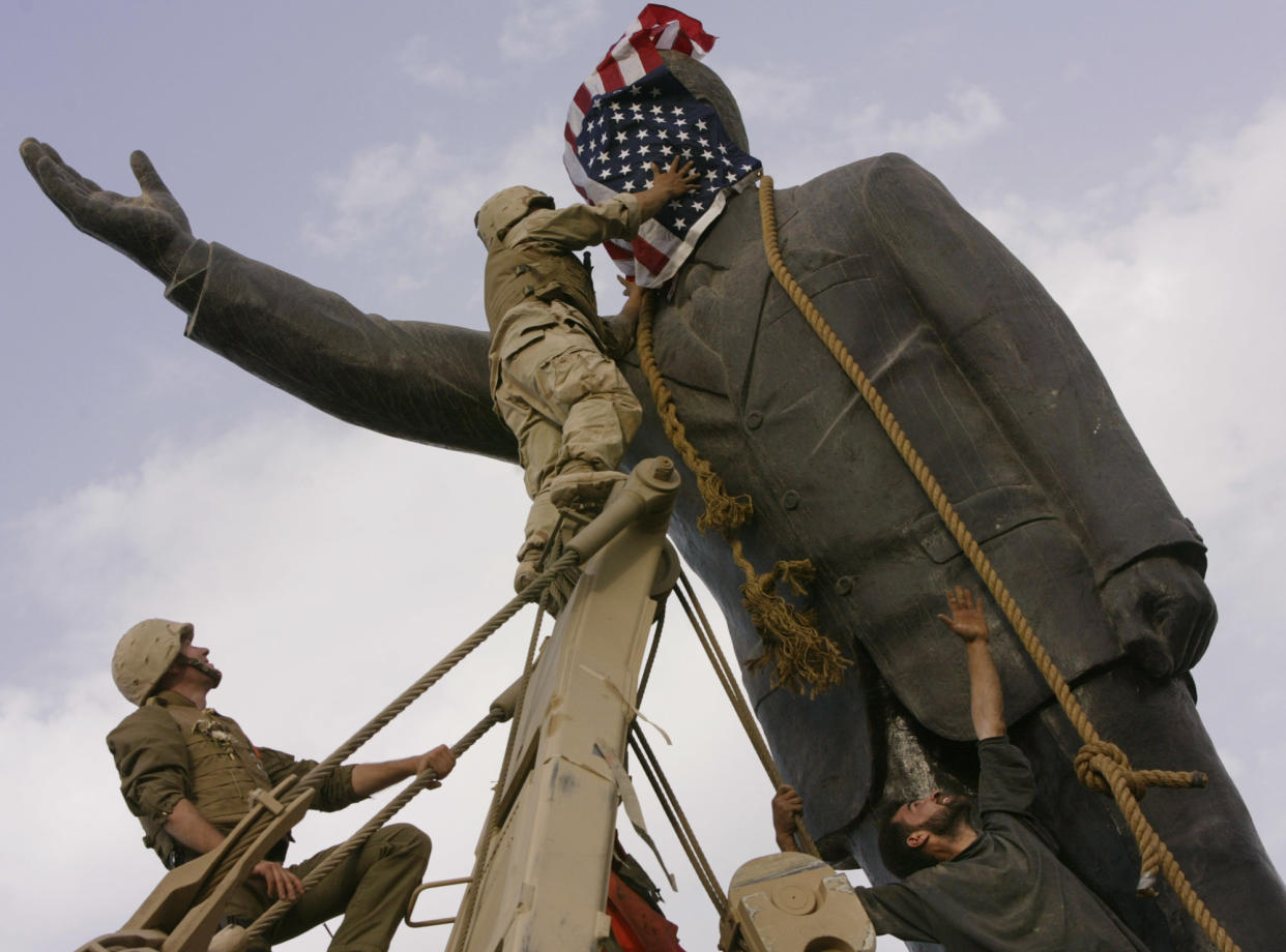 An Iraqi man, bottom right, watches Cpl. Edward Chin, on a ladder, cover the face of a statue of Saddam Hussein with an American flag, as another soldier holds a rope attached to the statue..