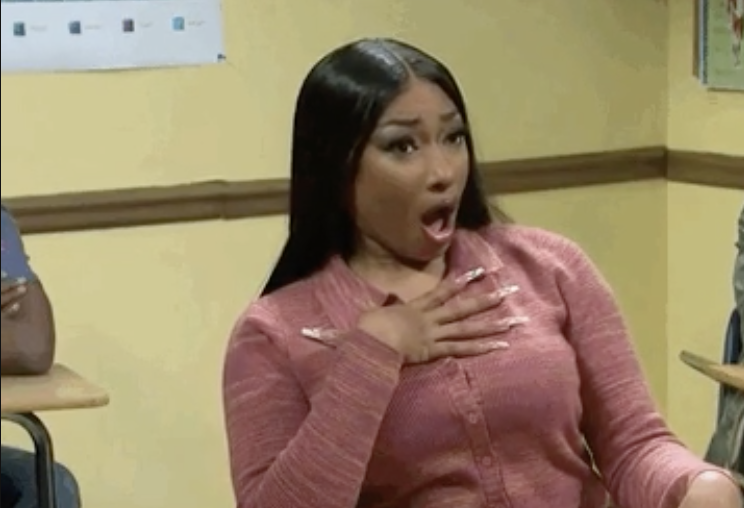 Nicki Minaj sitting at a desk in a classroom, looking surprised with her mouth open and a hand on her chest