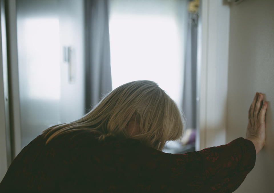 woman with blonde hair in sweater with hand on wall leaning in pain from nausea, dizziness, vomiting, tiredness, chest pain and headaches 