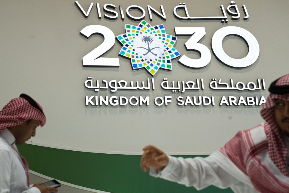 FILE - In this Sept. 10, 2019, file photo, men walk past a "Vision 2030" display at a stand about Saudi Arabia during the World Energy Congress in Abu Dhabi, United Arab Emirates. Lured by a long-looming stock offering of Saudi Arabia's massive state-run oil company, investors and business leaders have returned to the kingdom's capital for an investment forum overshadowed last year by the assassination of Washington Post columnist Jamal Khashoggi. (AP Photo/Jon Gambrell, File)