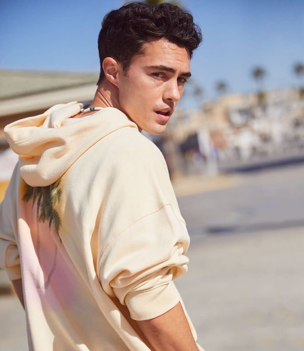 Darren Barnet wearing a Victoria's Secret apricot hoodie, back towards camera, he is slightly turned around looking at the camera, blurred background of a beach town