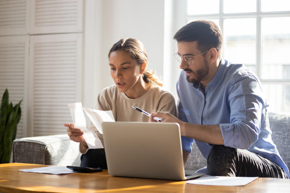 Concentrated young couple sit on couch busy managing finances paying bills on computer online, focused anxious millennial husband and wife calculate household expenses or taxes use internet banking