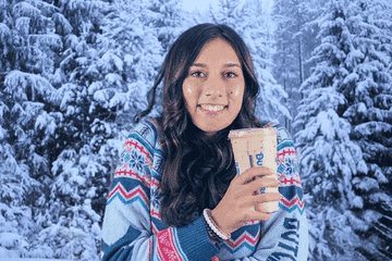 A person in a patterned sweater holds a drink, smiling in front of a snowy forest background