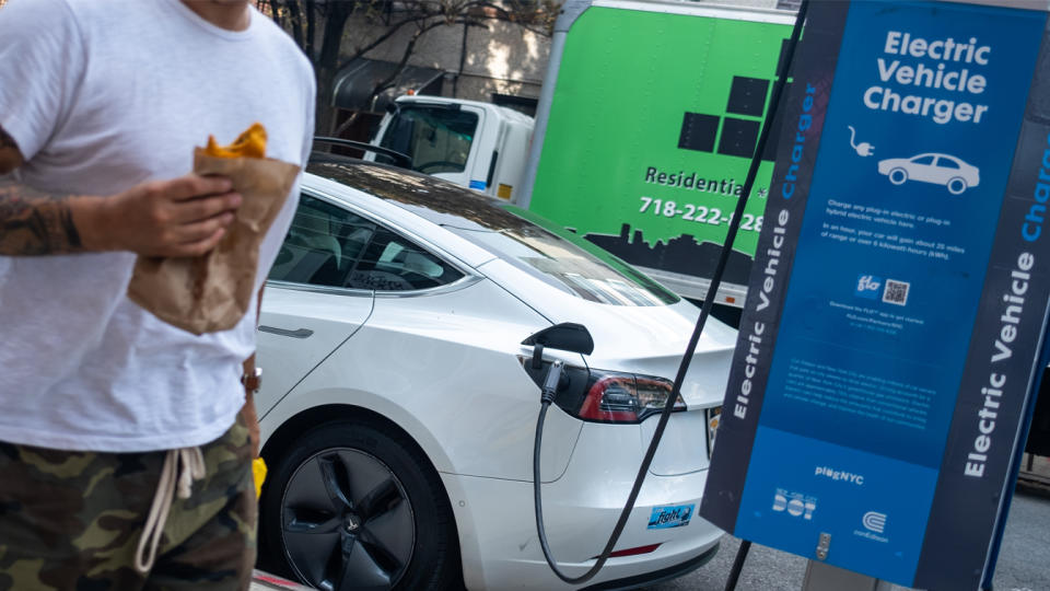 A person in a white T-shirt holding a brown bag containing what appears to be a partially eaten empanada walks past a Tesla charging at a public vehicle charger.