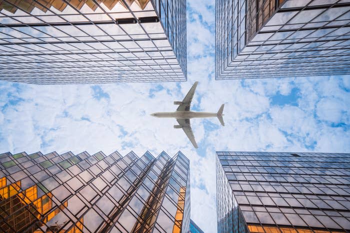 Looking up from the ground, a plane flies between high-rise buildings against a cloudy sky