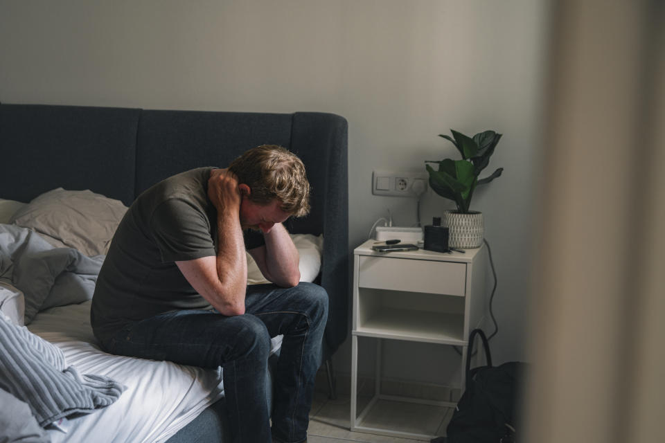 A depressed mature male sits on his bed. He clasps his hand behind his head