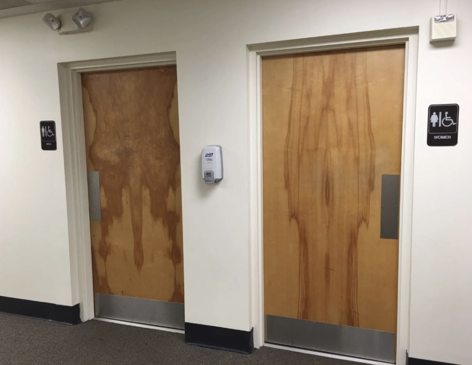 Two restroom doors in a hallway, with signs indicating accessible restrooms for men and women. A hand sanitizer dispenser is mounted between the doors