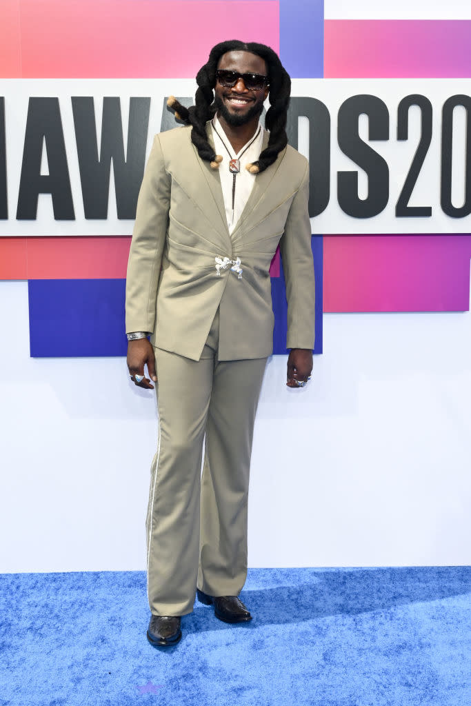 Shaboozey on the red carpet, wearing a stylish beige suit with a white shirt and unique accessories, smiles for the camera at an award event