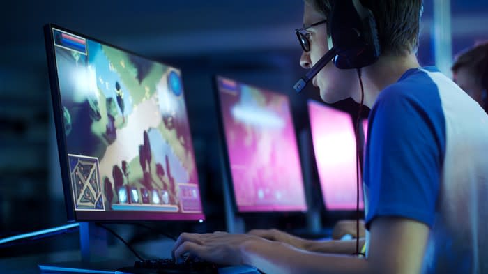 Young adult male playing a video game on a computer with three other computer screen and one other gamer next to him partially visible.