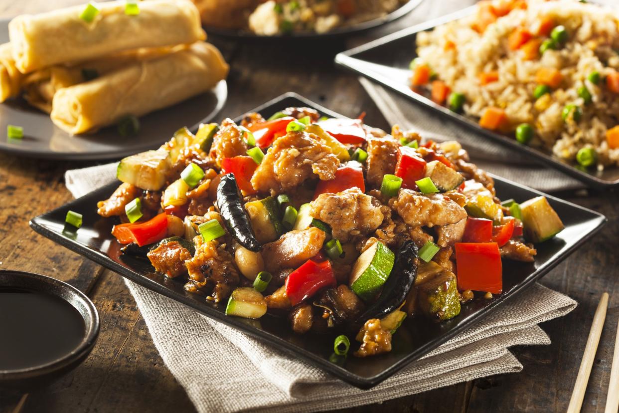 Easy kung pao chicken on a square black plate on a natural napkin with dipping sauce in a matching small bowl with eggrolls, stir-fried rice, and other Chinese foods in the background