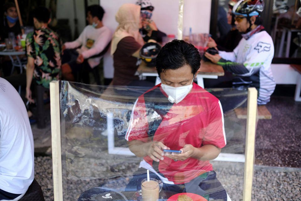 A man checks his mobile phone at a coffee shop, sitting in front of a plastic sheet barrier installed to help curb the new coronavirus outbreak in Makassar, South Sulawesi, Indonesia, Sunday, May 31, 2020. (AP Photo/Masyudi S. Firmansyah)
