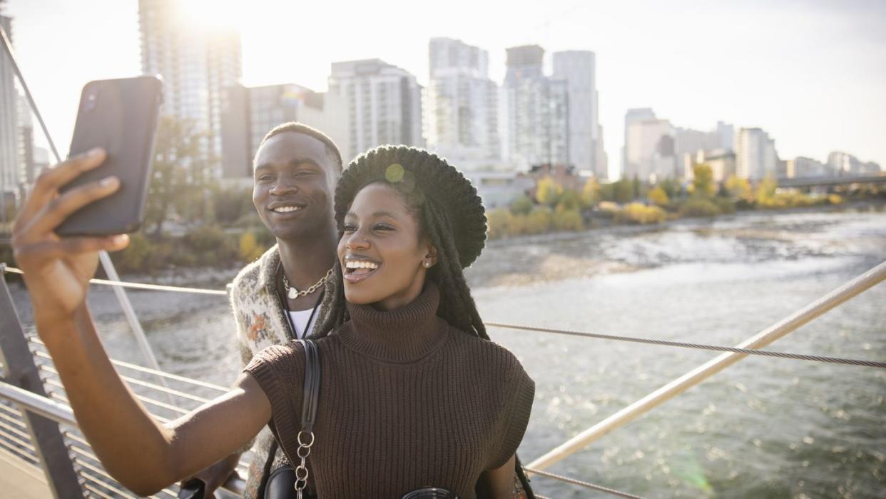 happy young couple taking selfie on sunny city footbridge over river