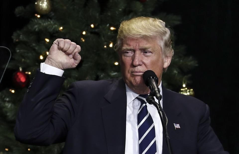 Donald Trump, vêtu d'un costume, fait un geste devant un podium avec un sapin de Noël en arrière-plan