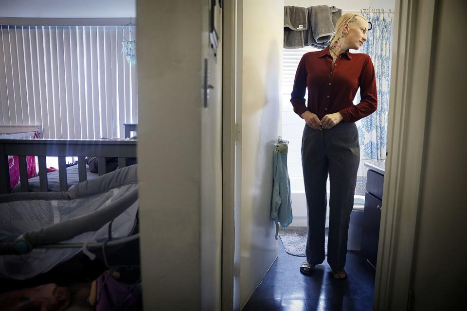 A woman in a red button-down shirt gets dressed in a bathroom and looks in the mirror.