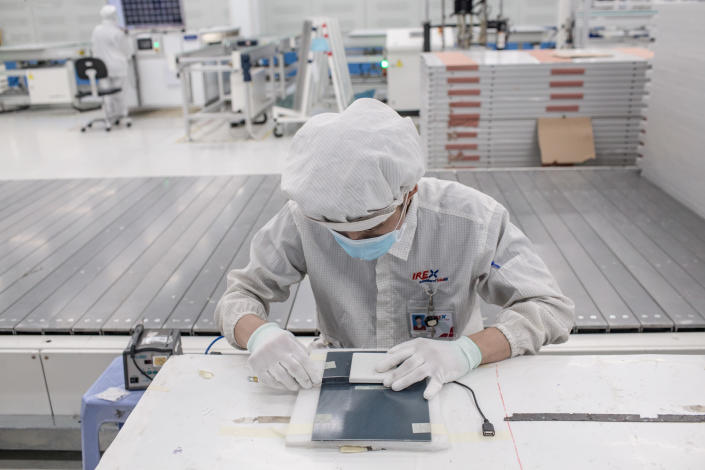 A worker wearing a mask, head covering and rubber gloves, leans over a solar battery to assemble it in a bare manufacturing facility, with one other worker visible in the distance. 