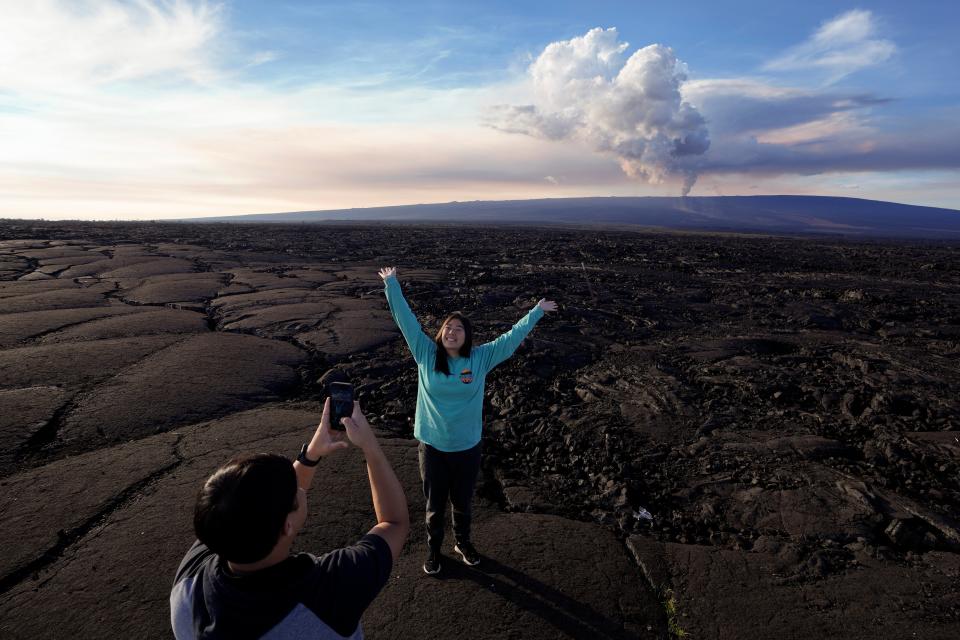 Kelly Ann Kobayashi raises her hands as she poses for a picture for Chad Saito, left, while standing on hardened lava rock from a previous eruption as the Mauna Loa volcano erupts, behind, Wednesday, Nov. 30, 2022, near Hilo, Hawaii (AP)