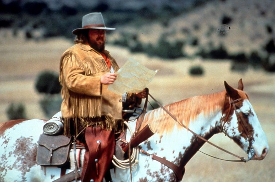 john candy wearing a cowboy costume, sitting on a horse and reading a map
