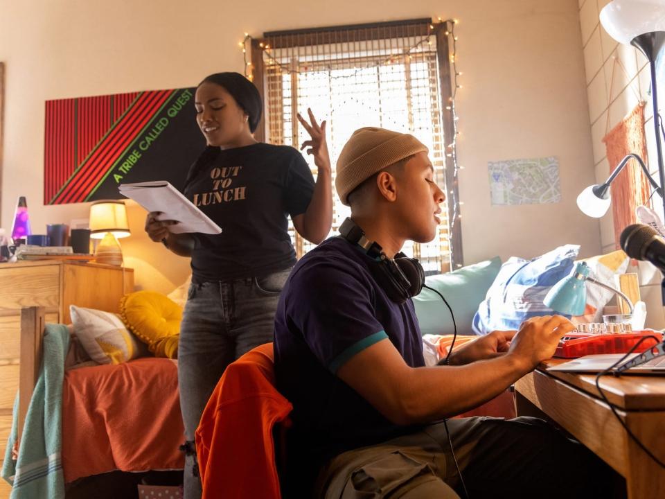 A woman rapping in a bedroom with a guy sitting at a desk in front of recording equipment.