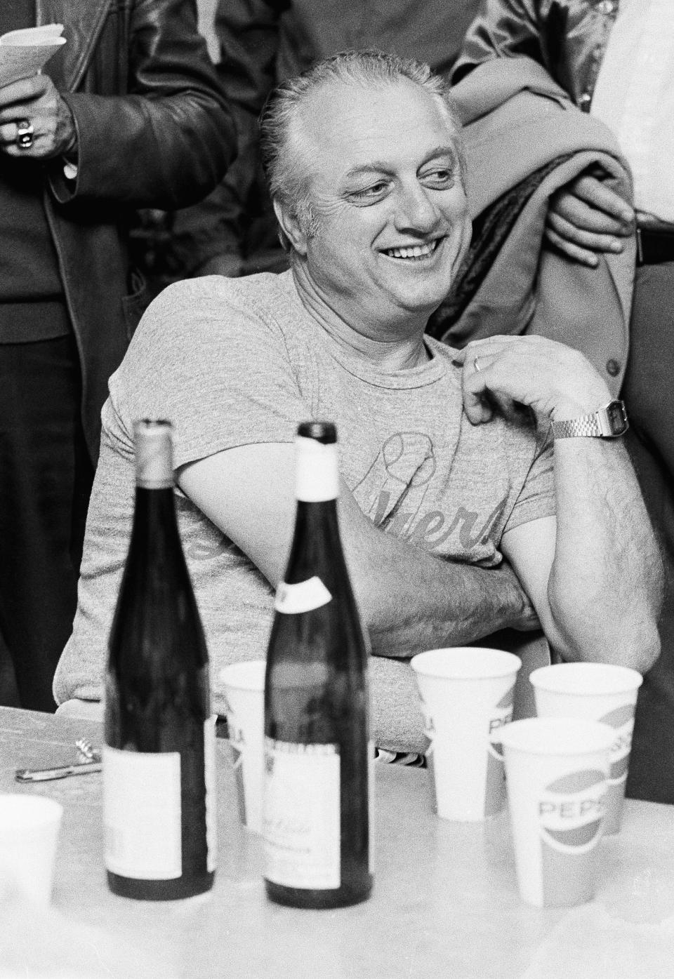 Tommy Lasorda grins as he answers a question during a champagne party in the Dodgers dressing room before their game with the Reds in Cincinnati on Tuesday, April 26, 1978. Lasorda invited newsmen in for the party as his answer to a new rule by the Reds barring visitors from their dressing room within 35 minute of game time. (AP Photo/ Brian Horton)