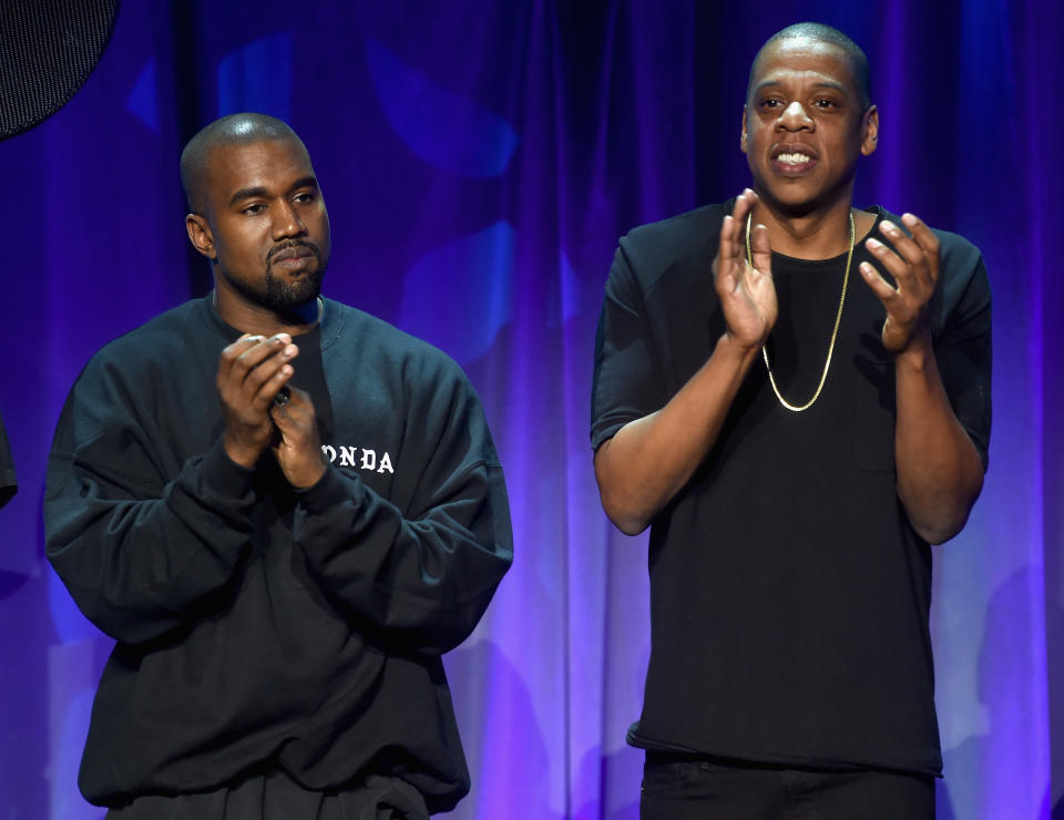 Two men on stage, one in a sweater and the other in a shirt, both applauding