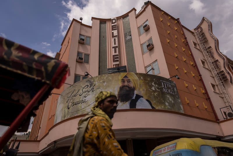 A rickshaw-puller stands in front of a poster of Aamir Khan-starrer "Laal Singh Chaddha", an official remake of the 1994 film "Forrest Gump" outside a cinema in New Delhi