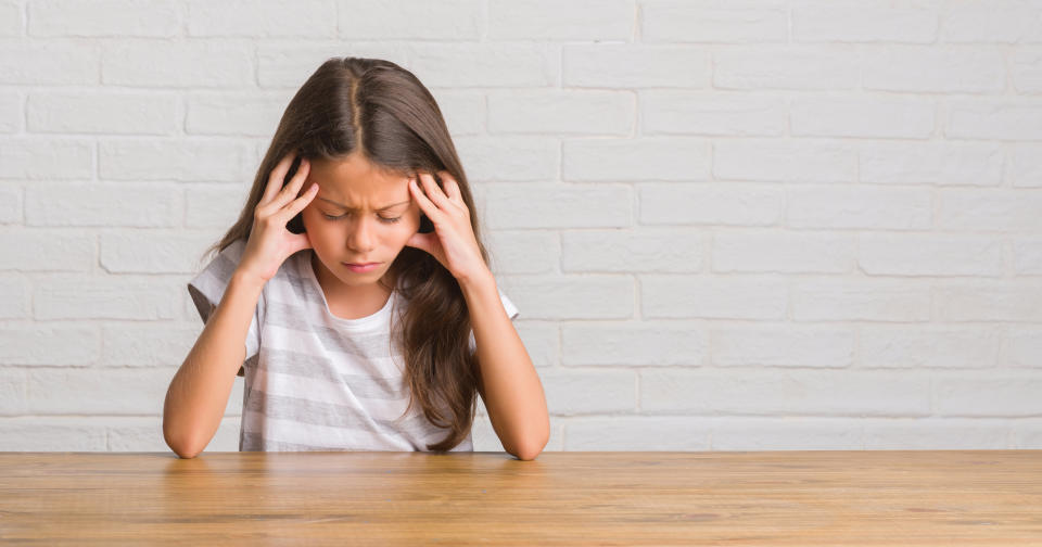 Young hispanic kid sitting on the table at home with hand on head for pain in head because stress. Suffering migraine.