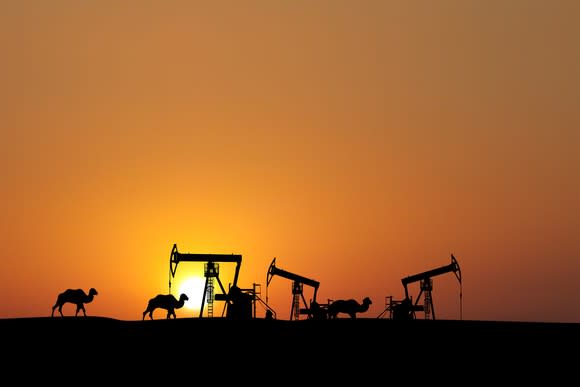 Camels walking past oil rigs at sunset.