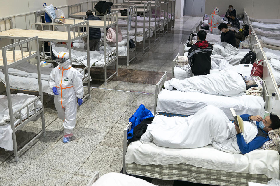 In this Wednesday, Feb. 5, 2020, photo, a medical worker in a protective suit walks by patients who diagnosed with the coronaviruses settle at a temporary hospital which transformed from an exhibition center in Wuhan in central China's Hubei province. Ten more people were sickened with a new virus aboard one of two quarantined cruise ships with some 5,400 passengers and crew aboard, health officials in Japan said Thursday, as China reported 73 more deaths and announced that the first group of patients were expected to start taking a new antiviral drug. (Chinatopix via AP)