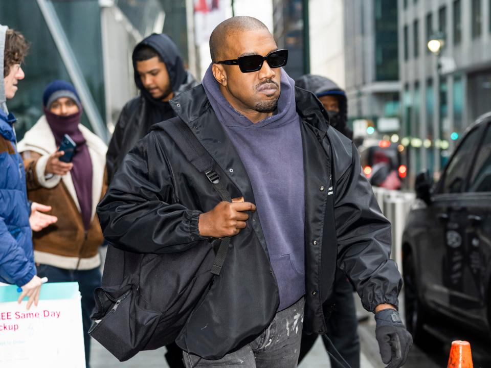 Kanye West walks down a sidewalk with a backpack slung over his shoulder