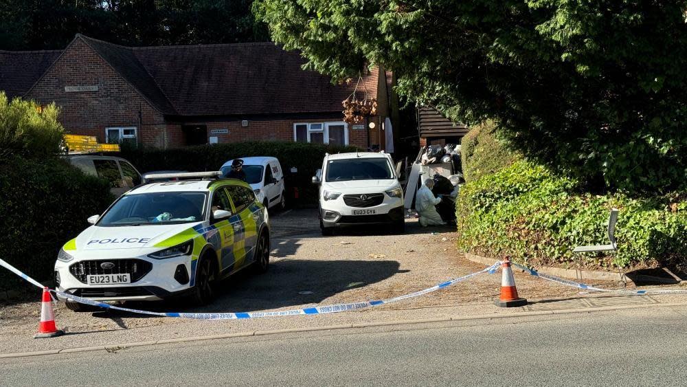 Four vehicles outside a property in Brentwood, showing a forensic officer in a white hazmat suit. There is also police tape and cones. 