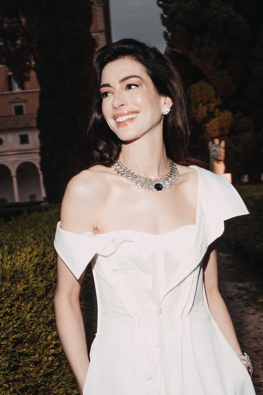 Anne Hathaway smiling, wearing an elegant off-the-shoulder white dress with a thigh-high slit and a statement necklace, standing outdoors near greenery