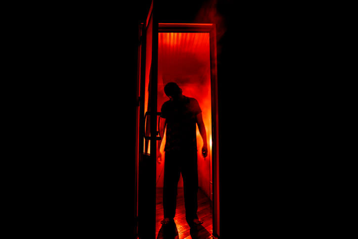 A silhouette of a person stands in a doorway with intense red lighting and smoke surrounding them. The figure appears mysterious and ominous against the dark background